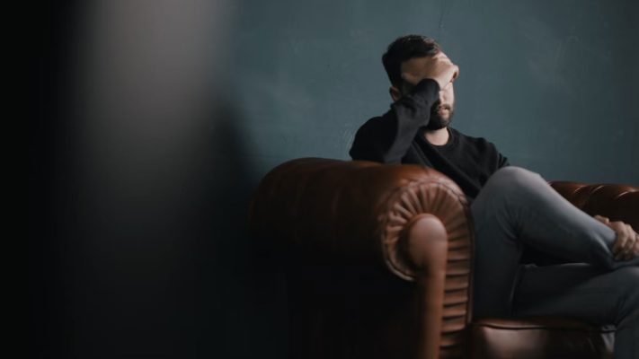 Male figure sitting on a brown leather chair holding his forehead with his right hand.